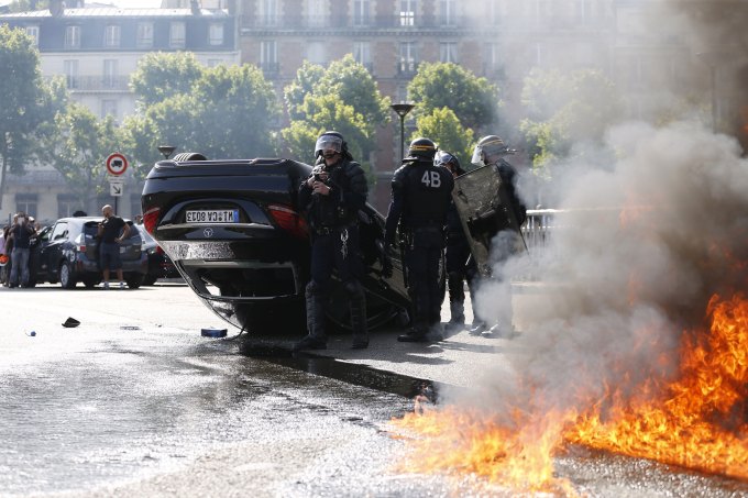 Smoke rises from a fire burning next to French CRS riot police standing near an overturned car as taxi drivers block Porte Maillot in Paris on June 25, 2015. Hundreds of taxi drivers converged on airports and other areas around the capital to demonstrate against UberPOP, a popular taxi app that is facing fierce opposition from traditional cabs. Access to three terminals at Paris-Charles de Gaulle airport and in a number of areas of Paris, especially Porte Maillot, were blocked. AFP PHOTO / THOMAS SAMSON (Photo credit should read THOMAS SAMSON/AFP/Getty Images)